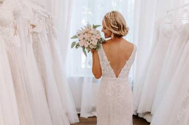 Braut steht im Brautmodenladen und zeigt den tiefen Rückausschnitt ihres Brautkleides.