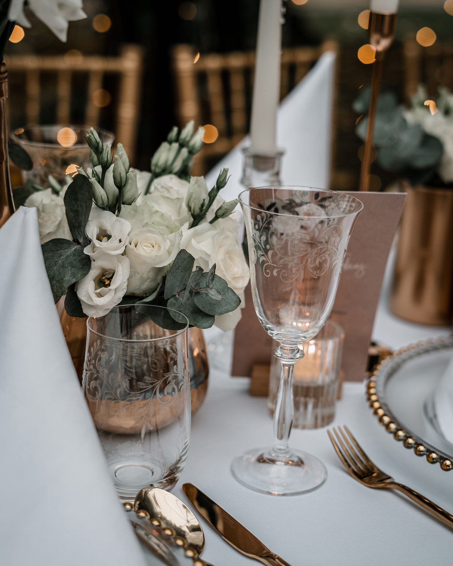 Der Hochzeitstisch wird in Detailaufnahme gezeigt: die durchsichtigen großen Teller mit Goldrand, goldenes Besteck und die Roségoldenen Vasen mit weißen Rosen und grünen Blättern geben ein schönes Bild ab. Die Papeterie passt ebenfalls zum Konzept.