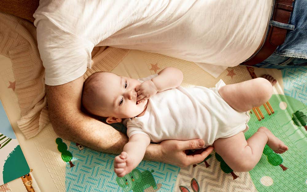 Baby liegt auf der bunten Spielematte in Papas Arm und hat die Hand im Mund