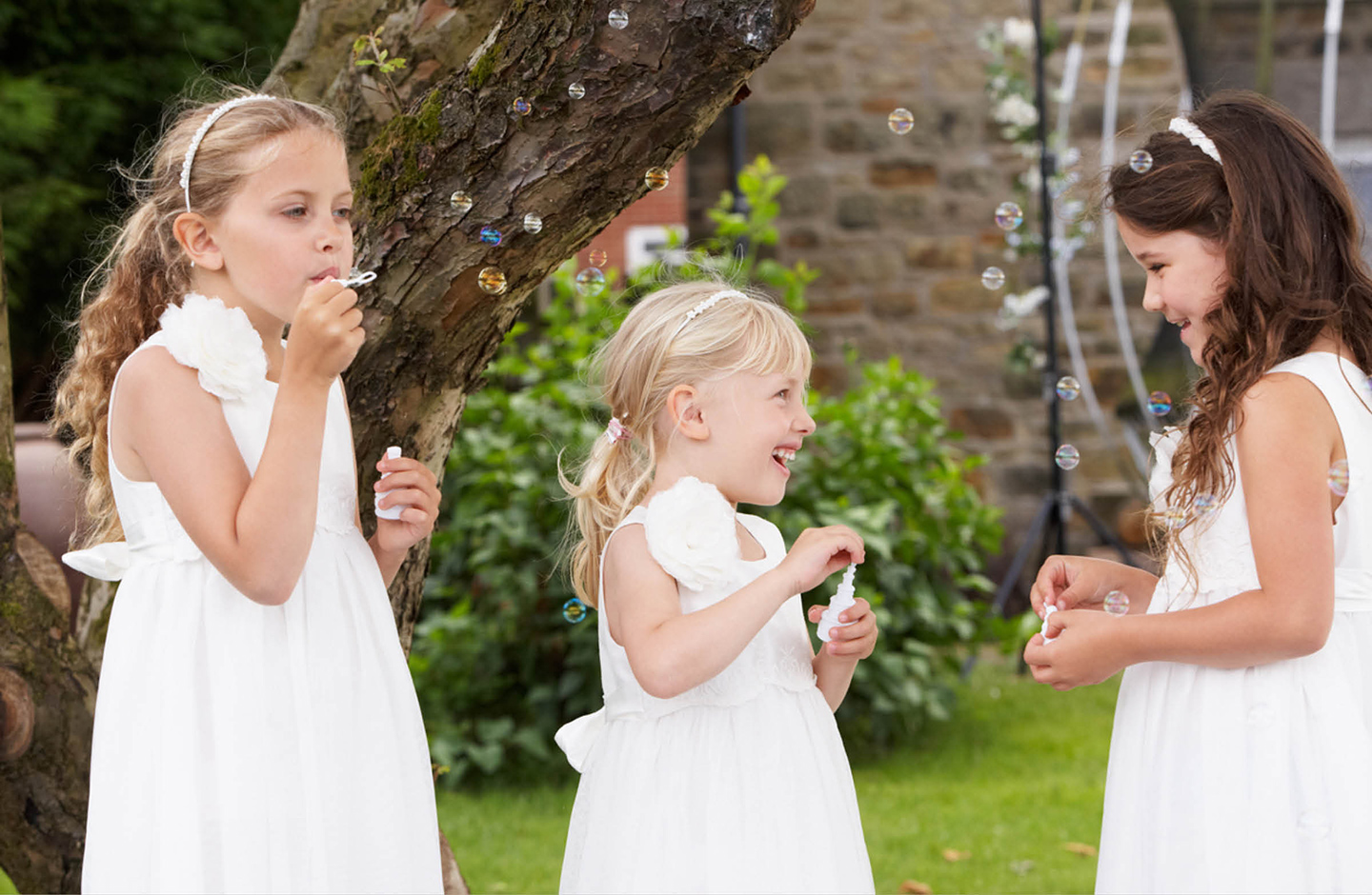 Drei kleine Mädchen, festlich in weißen Kleidern gekleidet, spielen im Garten mit Seifenblasen auf einer Hochzeitsfeier.