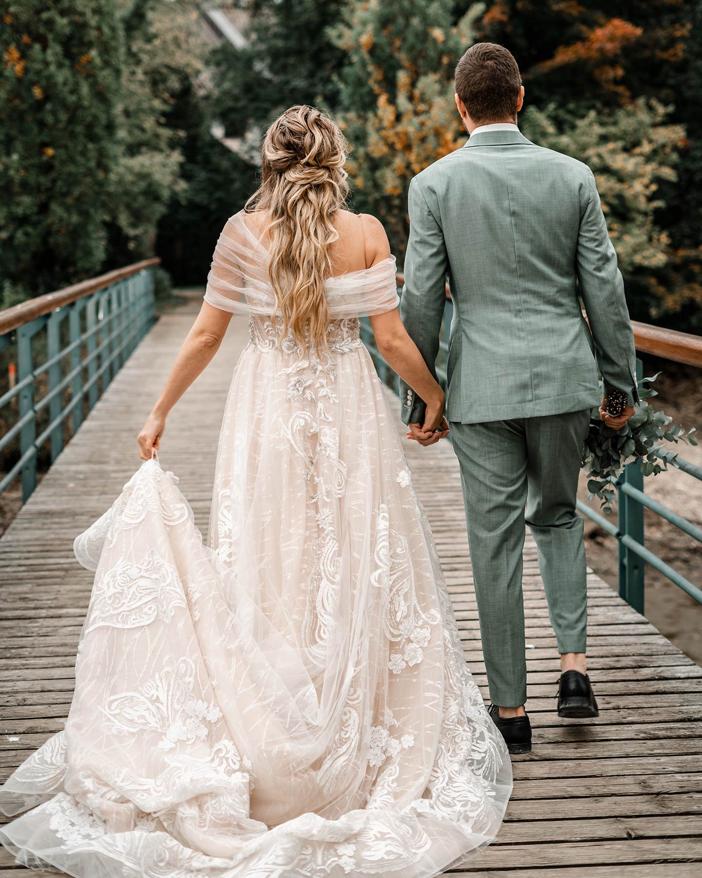 Das Hochzeitspaar ist von hinten zu sehen. Die Braut und der Bräutigam halten sich an der Hand und gehen gemeinsam über eine Holzbrücke.