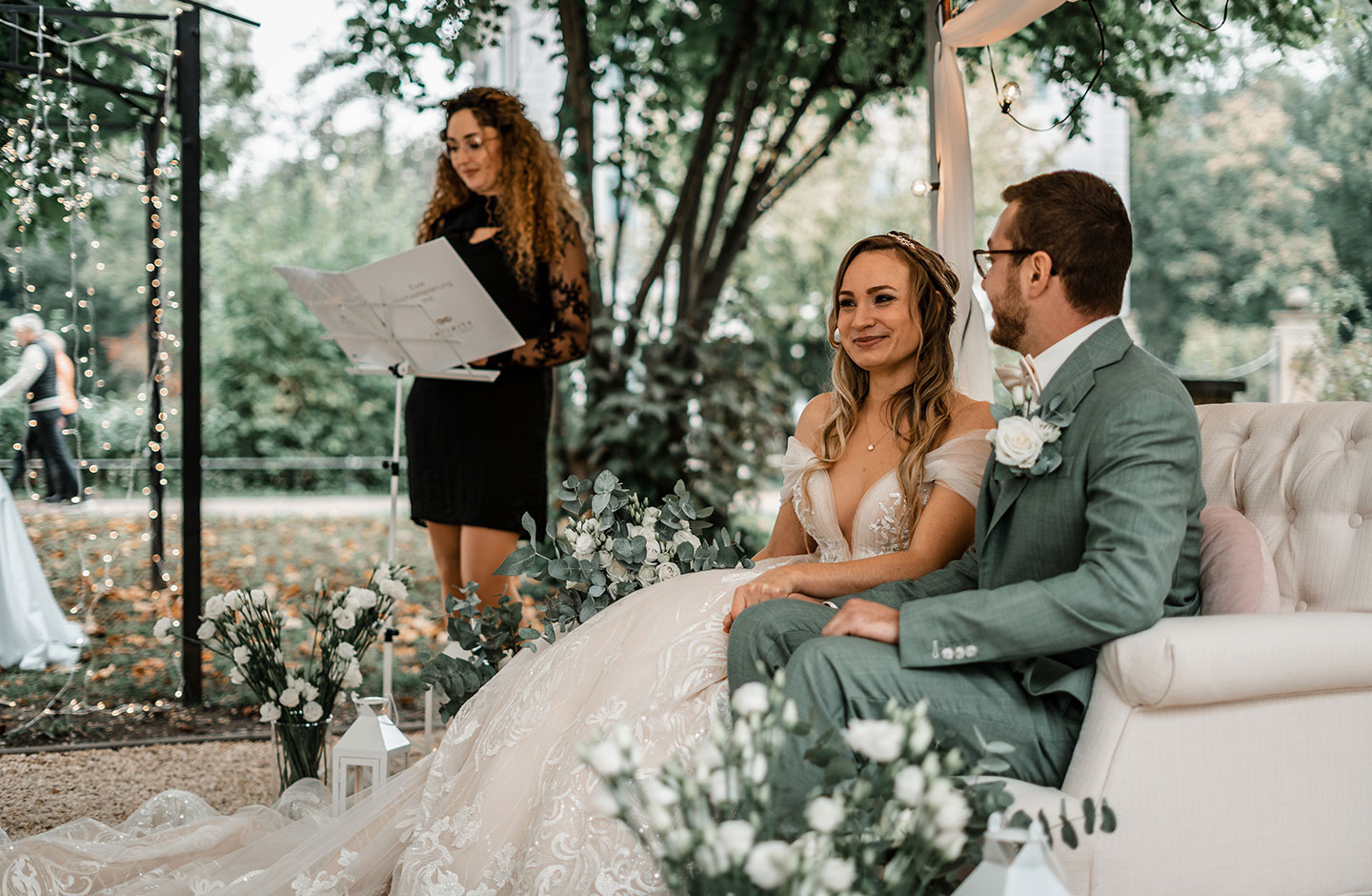 Das Hochzeitspaar bestehend aus der Braut im langen, cremefarbenen Tüllkleid und dem Bräutigam im grau-grünen Anzug, sitzen auf dem cremefarbenen Sofa unter dem Traubogen und sind bereit für die Trauzeremonie. Im Hintergrund ist die Traurednerin zu sehen.