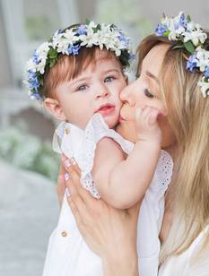 Mama mit Blumenkranz im Haar küsst ihr Baby am Tag der Taufe liebevoll auf die Wange.