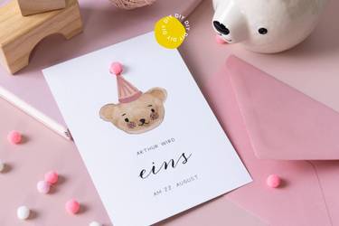 Einladungskarte zum 1. Geburtstag mit Bär-Illustration liegt auf einem rosa Umschlag.