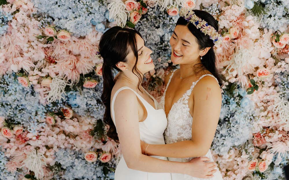 Verliebtes Hochzeitspaar hält sich in den Armen vor einer Blumenwand mit rosa und blauen Blumen