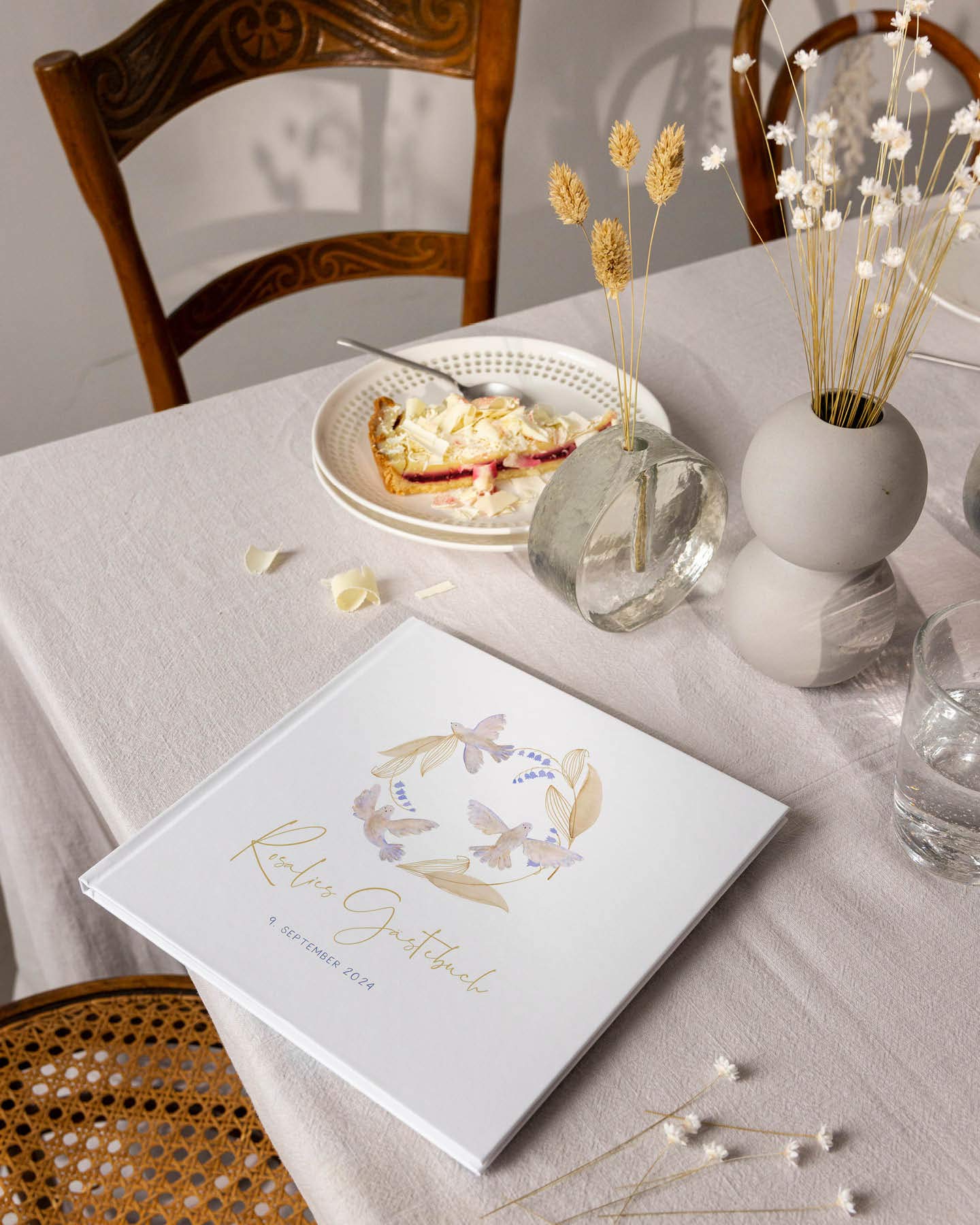 Personalisiertes Gästebuch mit zarten Tauben-Illustrationen liegt auf einem Tisch mit heller Leinentischdecke zum Namensgebungsfest.