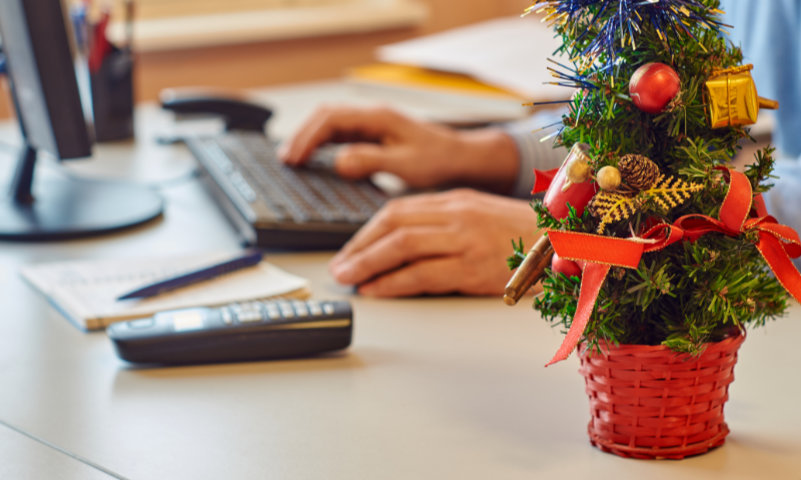 Am Schreibtisch sitzend vor dem Computer befindet sich ein Mitarbeiter eines Unternehmens. Sein Tisch ist geschmückt mit weihnachtlichen Accessoires. Zu dieser Zeit machen sich viele Unternehmer Gedanken, wie sie trotz DSGVO Weihnachtskarten verschicken