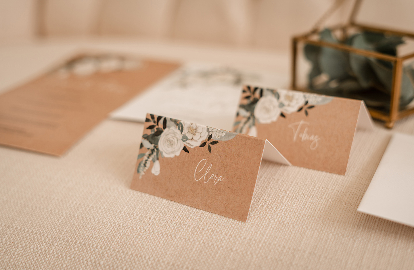  Die Hochzeitspapeterie wurde auf dem cremefarbenen Sofa für ein Foto platziert. Die im Kraftpapier-Look mit den Namen der Gäste beschrifteten Tischkärtchen greifen das Design der weißen Blüten und grünen Blätter auf.