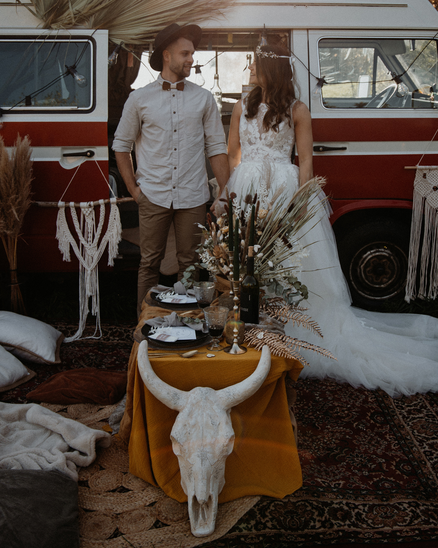 Verliebtes Boho Brautpaar feiert im Picknick Hochzeit. Als Dekoration werden ein Bulli, Pampasgras und Makramee-Elemente verwendet.