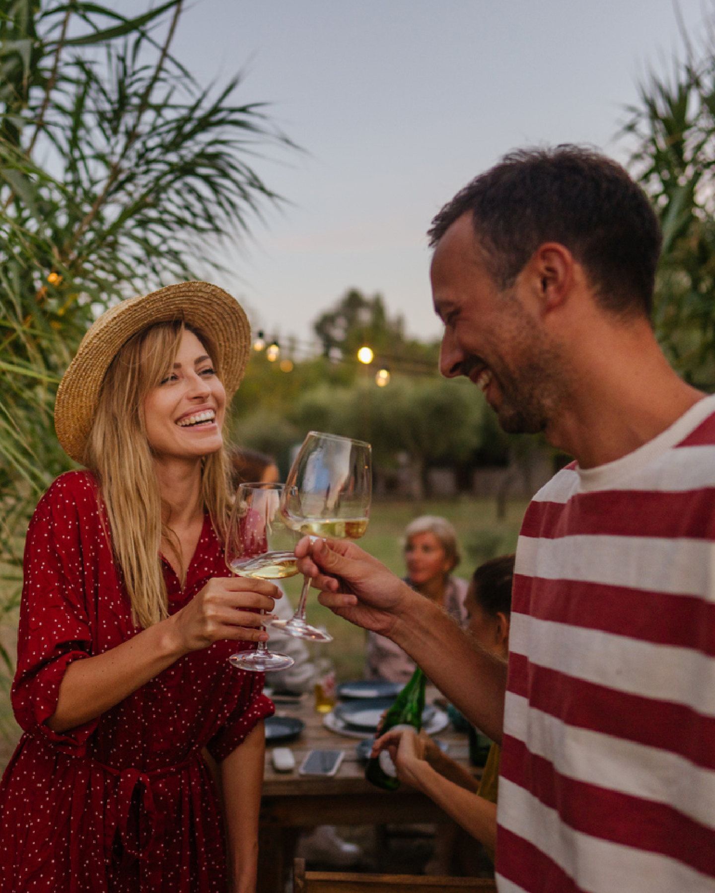 Frau in rotem Kleid mit Strohhut und Mann in rot-weiß restreiftem TäShirt stoßen bei Gartenparty mit Weißwein an