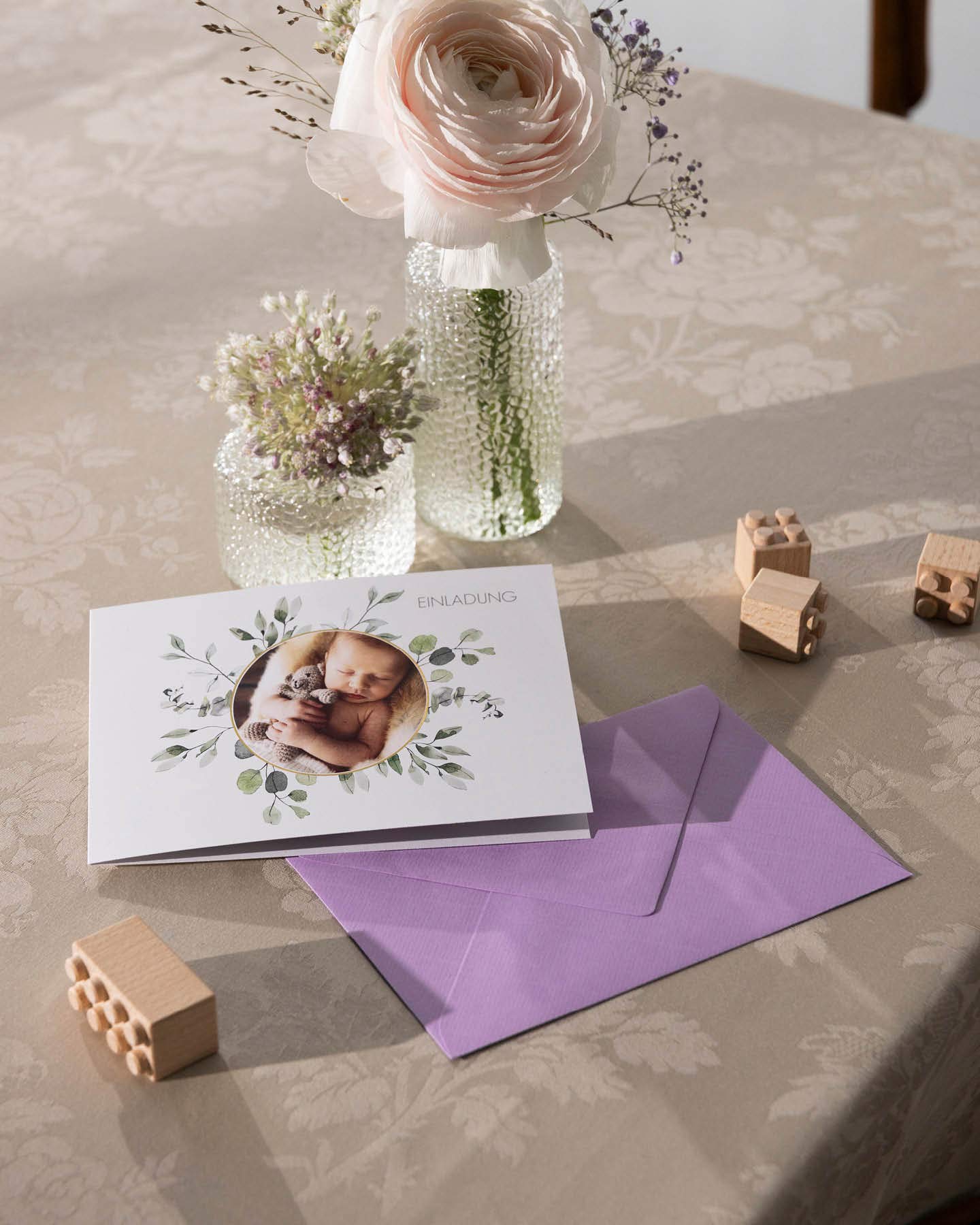 Einladungskarte zur NAmensgebungsfeier mit Umschlag auf einem Tisch mit  zwei Glasvasen und Holzspielzeug