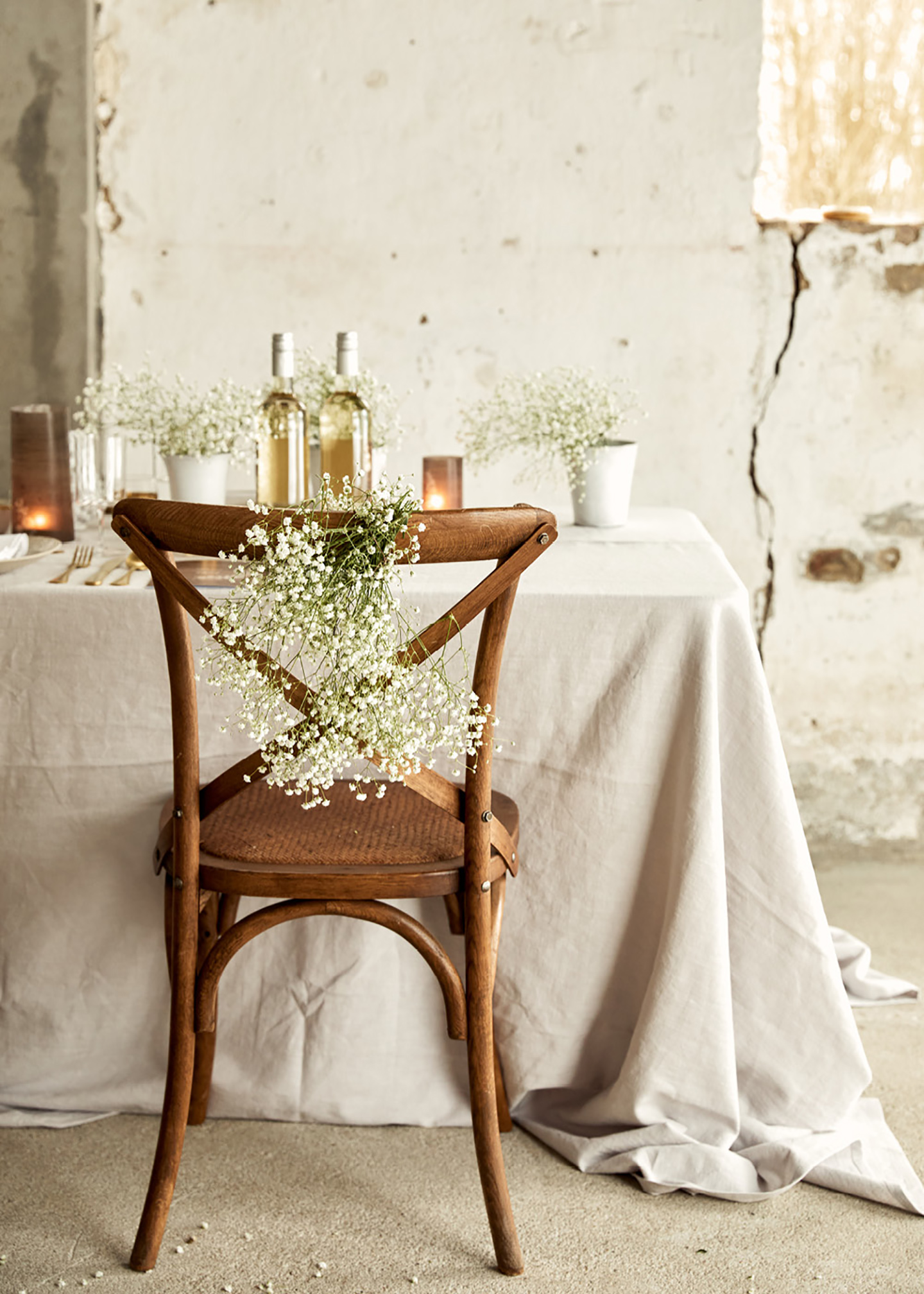 Tischdeko Hochzeit: Eine rustikale Location im Shabby-Chic-Look, ein im Vintage Stil dekorierter Hochzeitstisch mit weißer Tischdecke und dunklen Stühlen sowie goldenes Besteck und Schleierkraut geben ein harmonisches Bild.