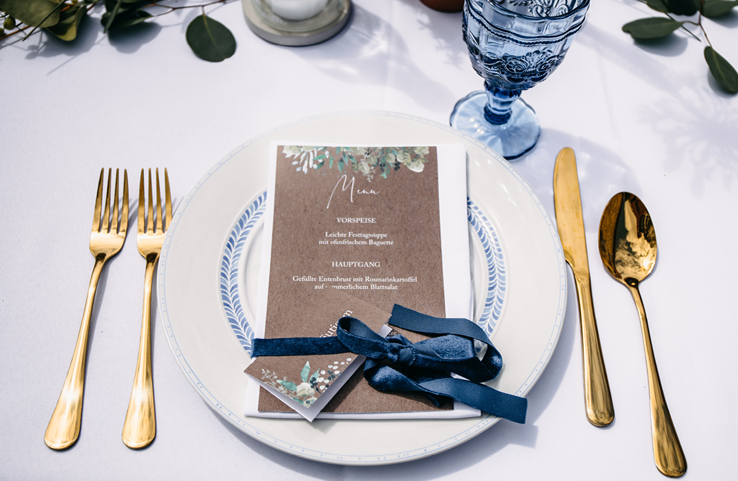 Menükarte & Namenskarte aus Kraftpapier mit Greenery Illustrationen liegen auf einem weißen Teller auf der Hochzeitstafel.