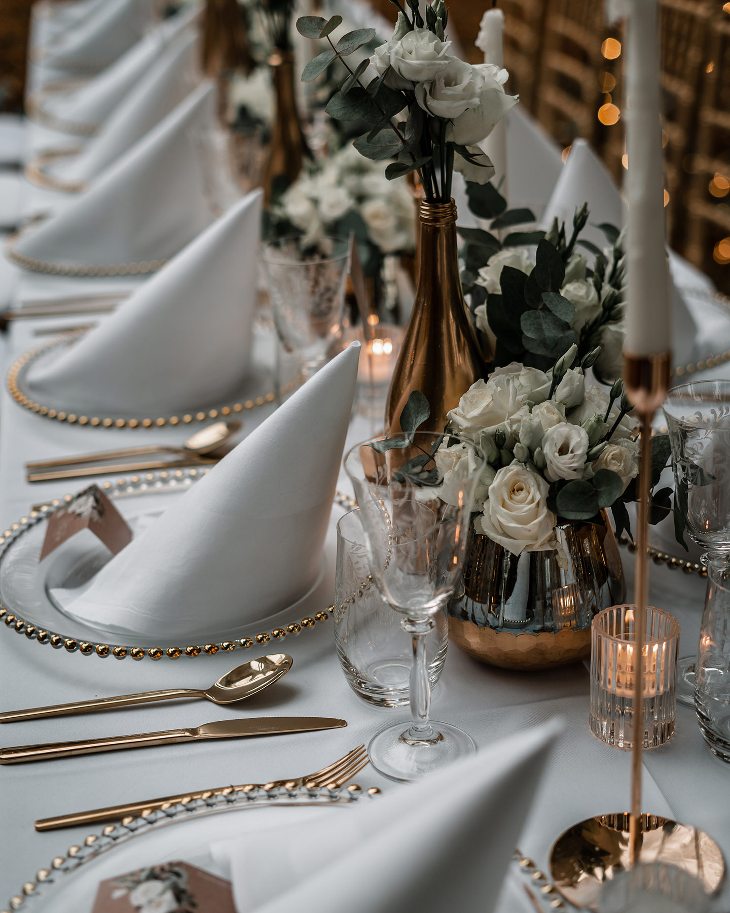 Der Hochzeitstisch wird in Detailaufnahme gezeigt: die durchsichtigen großen Teller mit Goldrand, goldenes Besteck und die Roségoldenen Vasen mit weißen Rosen und grünen Blättern geben ein schönes Bild ab.