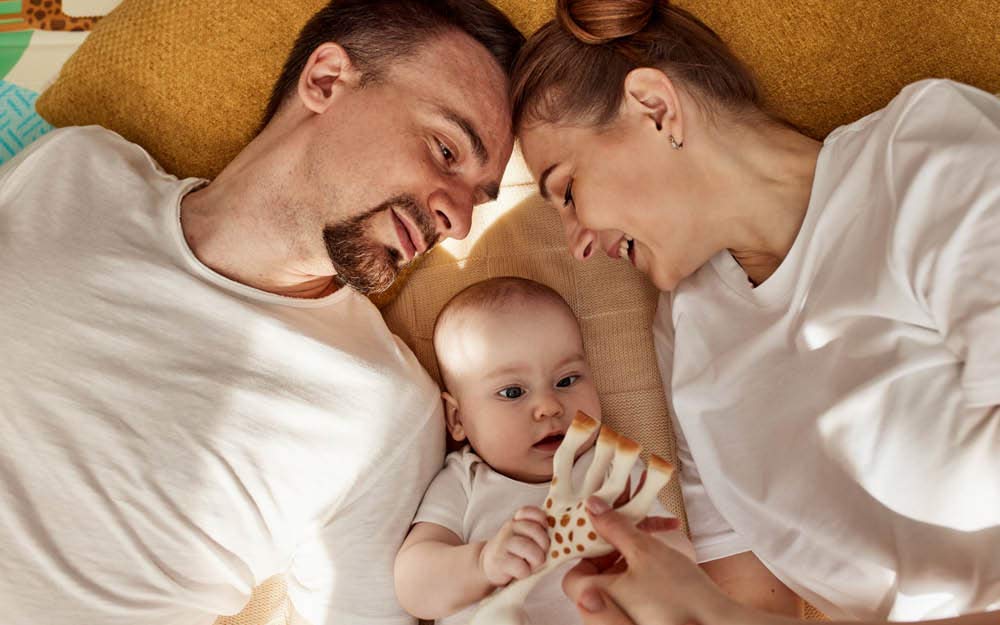Mama und Papa kuscheln liegend mit Baby, während dieses ein Babyspielzeug hält