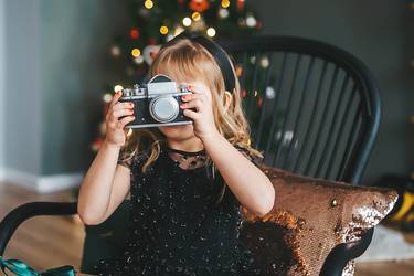 Ein kleines Mädchen sitzt in einem festlichen Kleid auf einem Schaukelstuhl. In der Hand hält sie eine Kamera. Im Hintergrund glänzt der geschmückte Weihnachtsbaum.