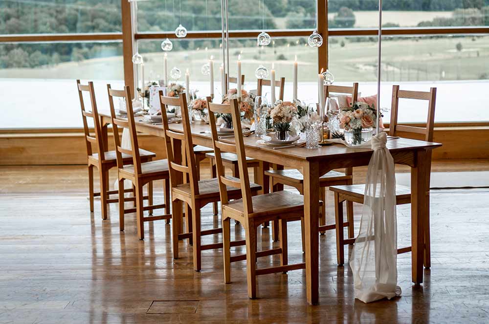 Hochzeitstafel: Ein großer Holztisch ist dekoriert mit Blumen in Koralle, weißen Kerzen und Teelichtern aus Glas.