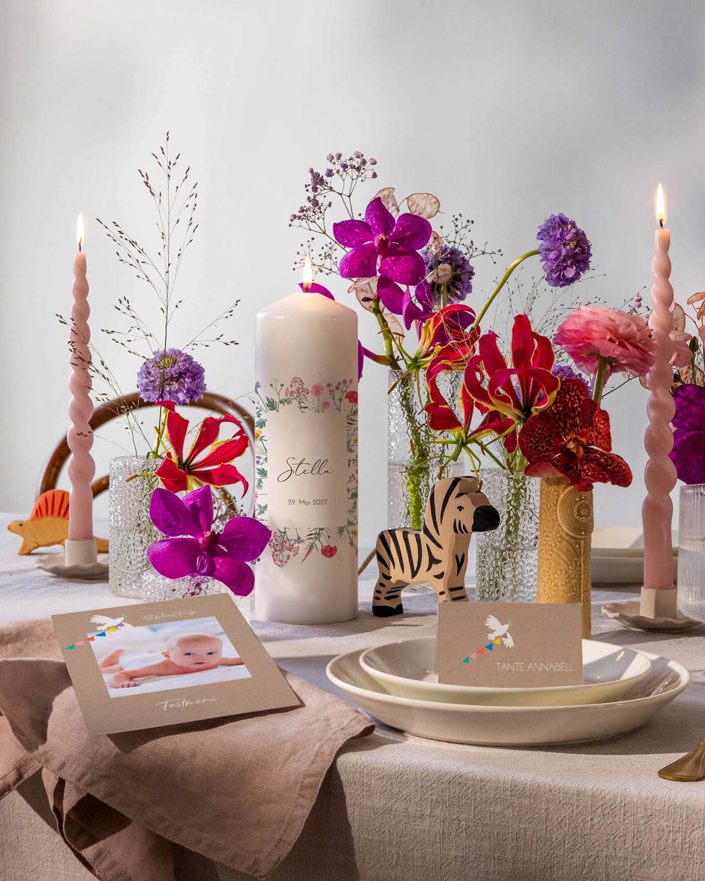 Die Festtafel zur Namensgebungsfeier ist mit roten, violetten und pinken Blumen dekoriert. Eine Lebenskerze, eine Menükarte und eine Namenskarte runden den Sitzplatz der Gäste ab.