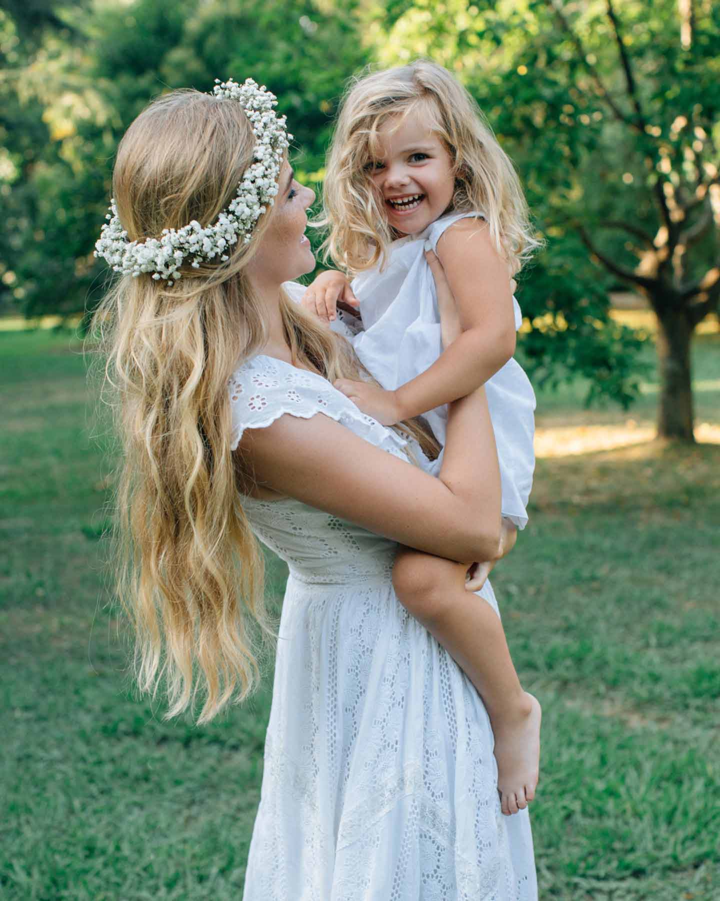 Mutter im weißen Kleid und mit Blumenkranz im Haar, hält ihre Tochter lachend im Arm am Tag der Namensgebungsfeier