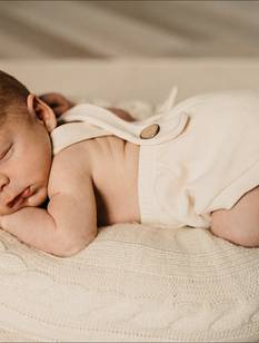 Niedliches Baby in Latzshorts schläft bäuchlings auf einem cremefarbenen Kissen Kissen