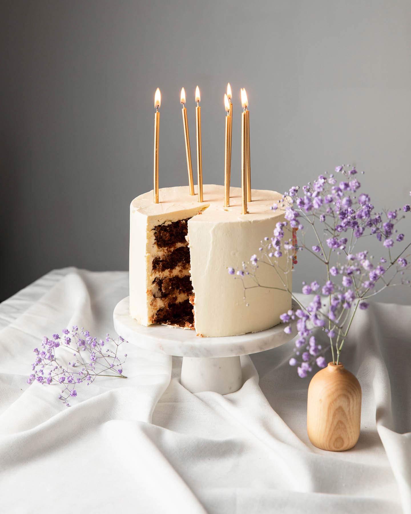 Weiße Torte mit Schoko-Füllung steht auf einer Mamorplatte auf einem Tisch. Goldene Kerzen brennen und stecken in der Geburtstagstorte.