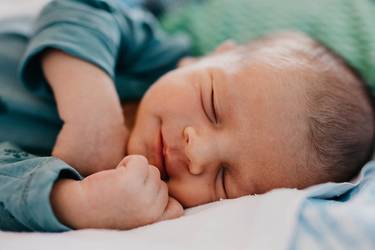 schlafendes Baby liegt auf der Seite und trägt einen blauen Body. Seine Hände sind zu einer Faust.