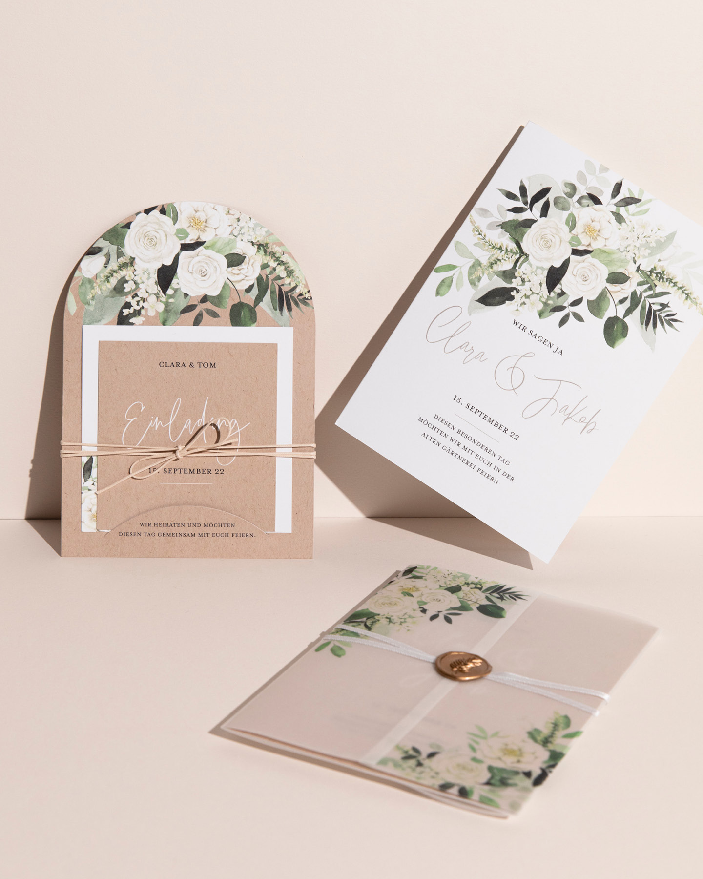 Drei Hochzeitseinladungen mit Greenery & weißen Blumen Illustrationen lehnen an hellem Hintergrund.