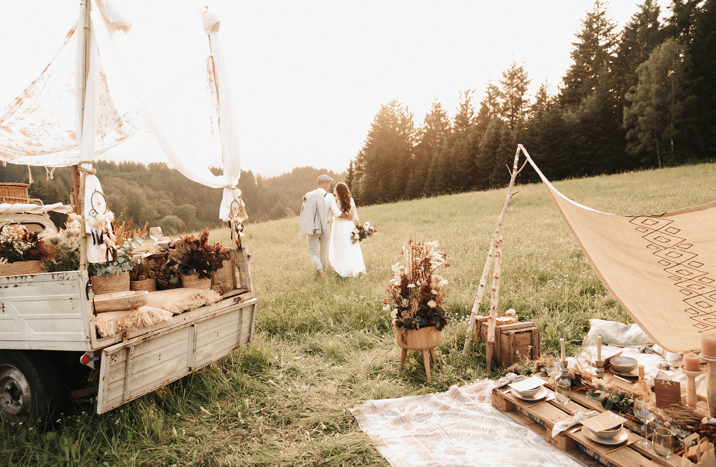 Brautpaar läuft Hand in Hand über Wiese Richtung Sonnenuntergang. Im Vordergrund ist ihr Boho Picknick sichtbar.