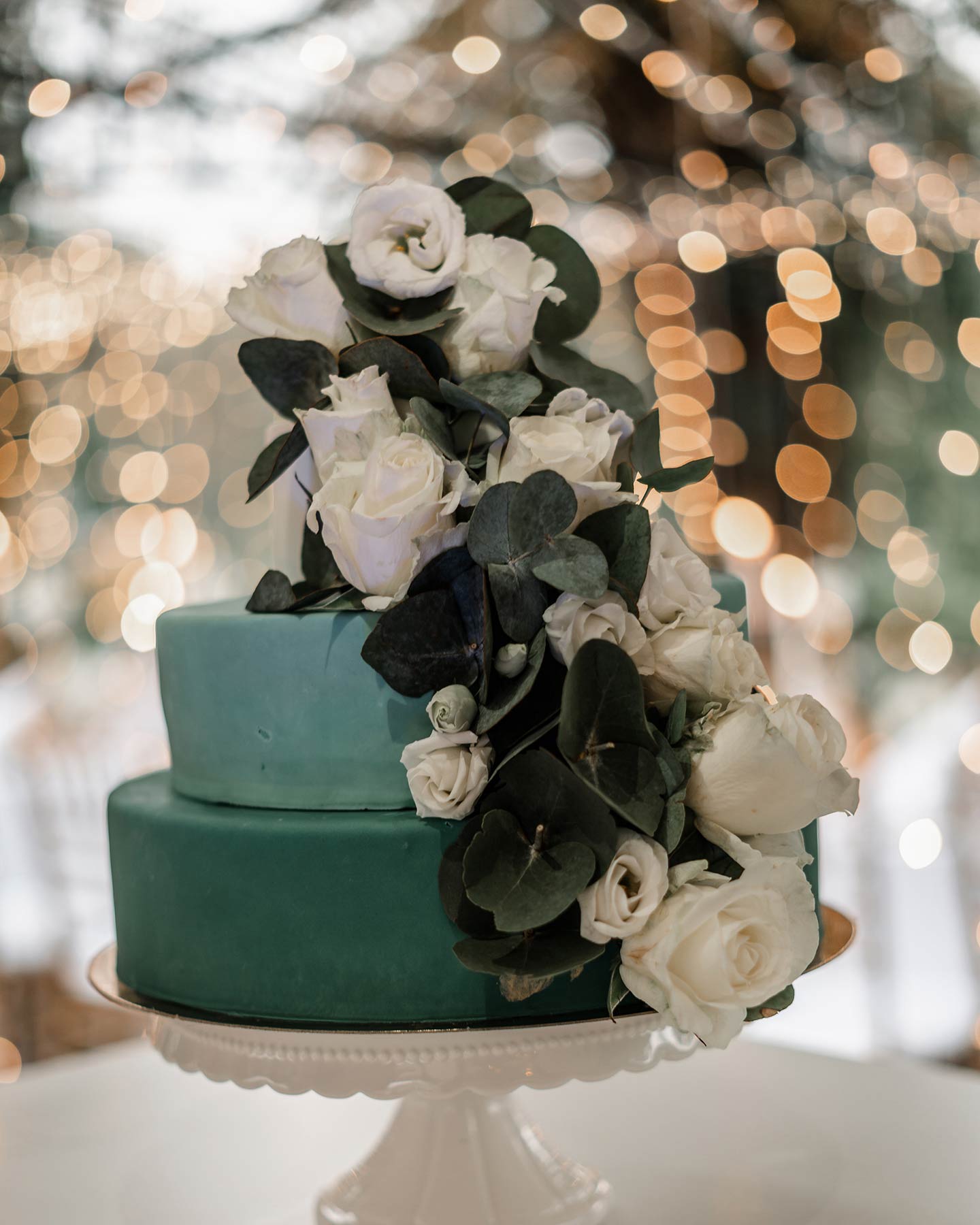 Die Hochzeitstorte ist in Gänze zu sehen: sie besteht aus zwei Stöcken in Grün-Türkistönen, geschmückt mit Blüten in weiß und grünen Blättern. Im Hintergrund ist das Lichtermeer der Lichterketten zu sehen.