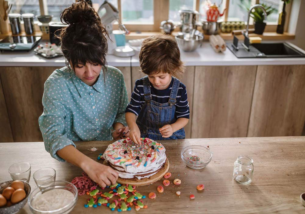 Mama backt mit ihrem Kind zusammen einen Geburtstagskuchen und dekoriert ihn mit Süßigkeiten.