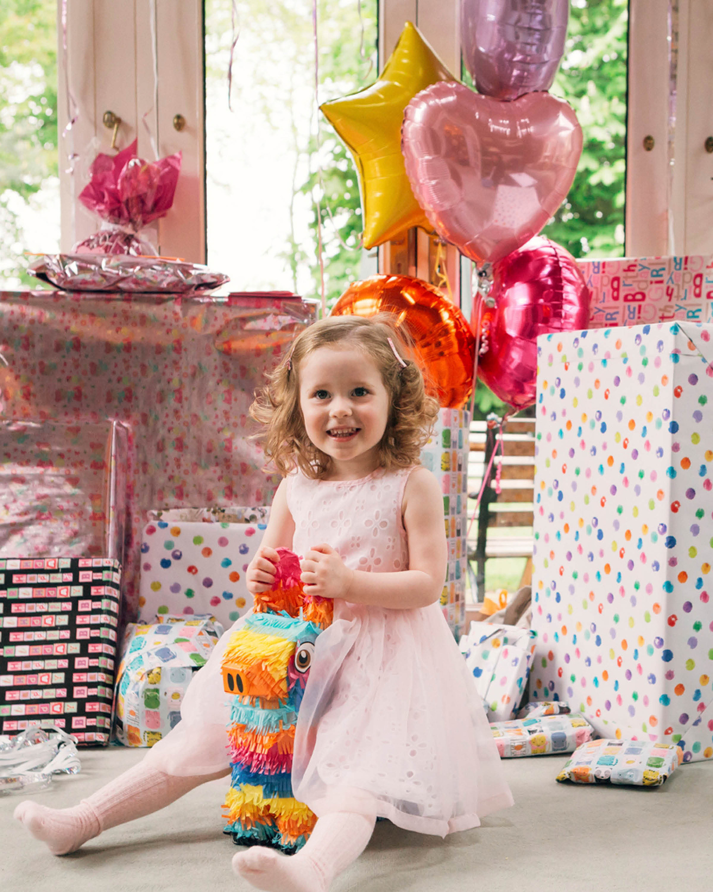 Mädchen in rosafarbenem Kleid sitzt vor einem großen Berg an Geburtstagsgeschenken. Helium gefüllte Luftballons in Herz- und Sternform zieren die Geschenke.