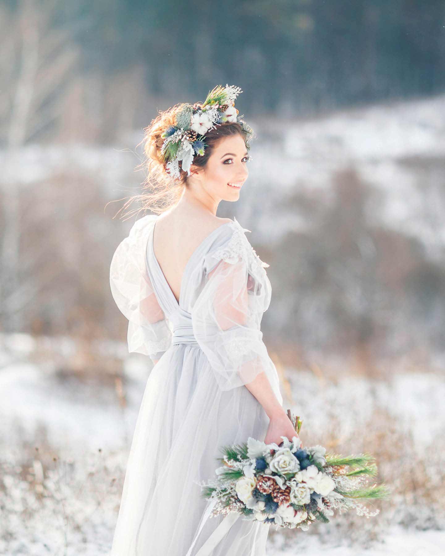 Braut steht vor verschneiter Winterlandschaft mit Braustrauß in der Hand